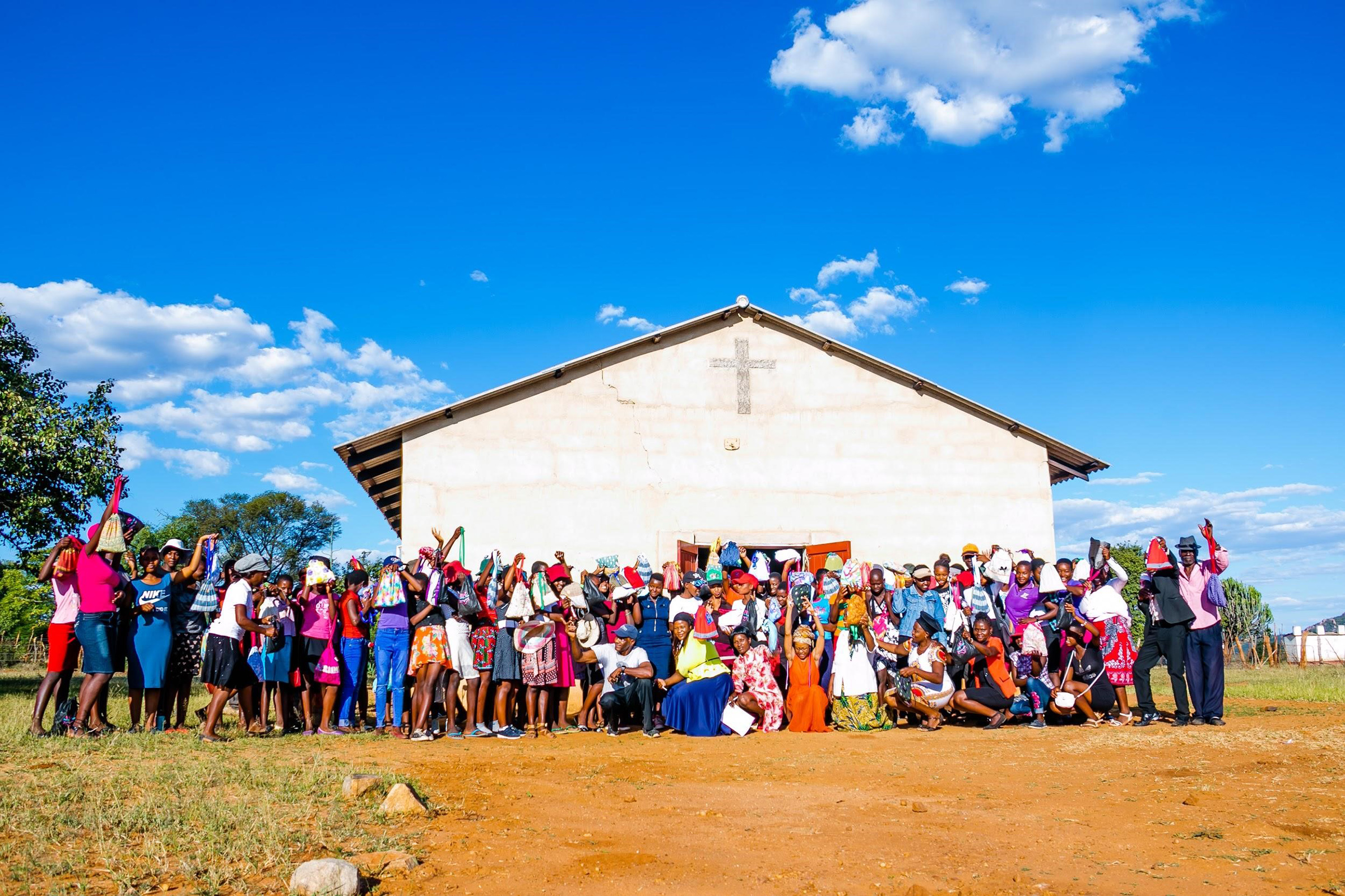A large group of Mbizingwe people stand in front of a large white building holding up bags.