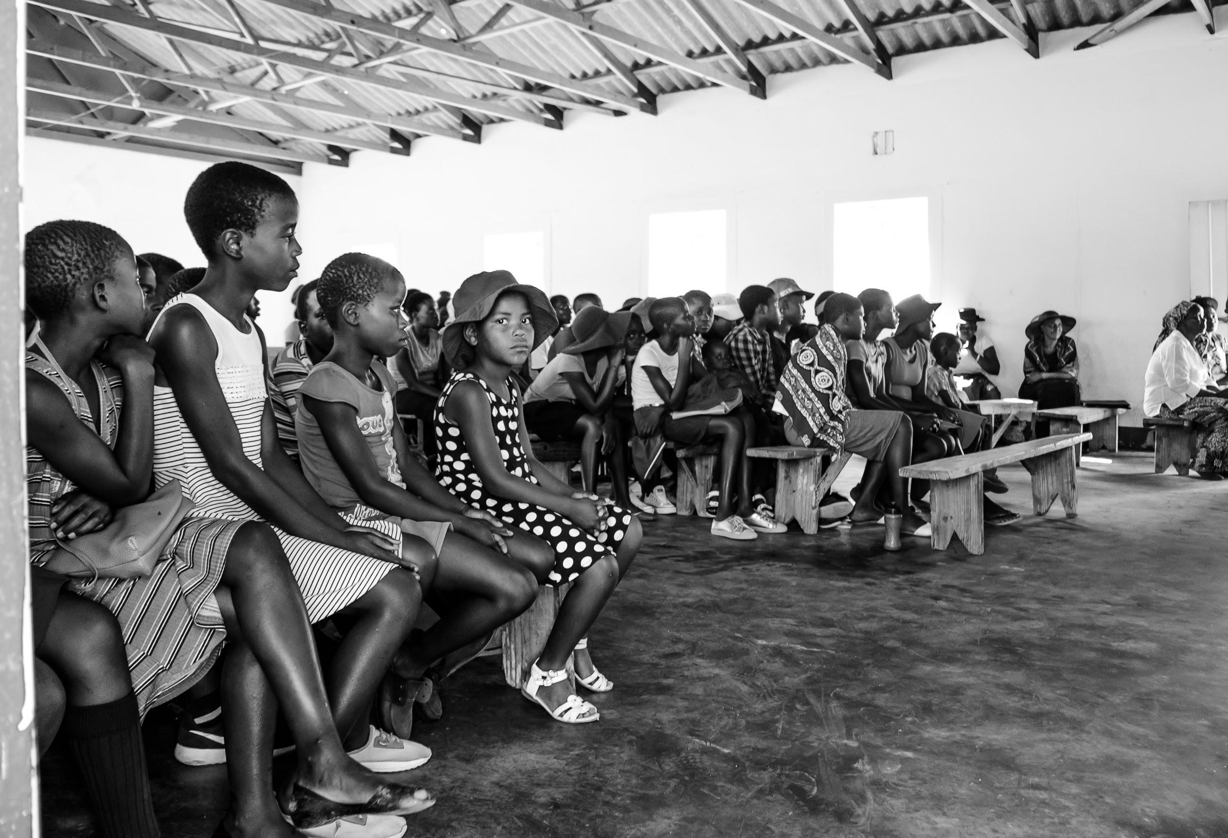 A large group of African children in a large building; one child is looking at the camera.