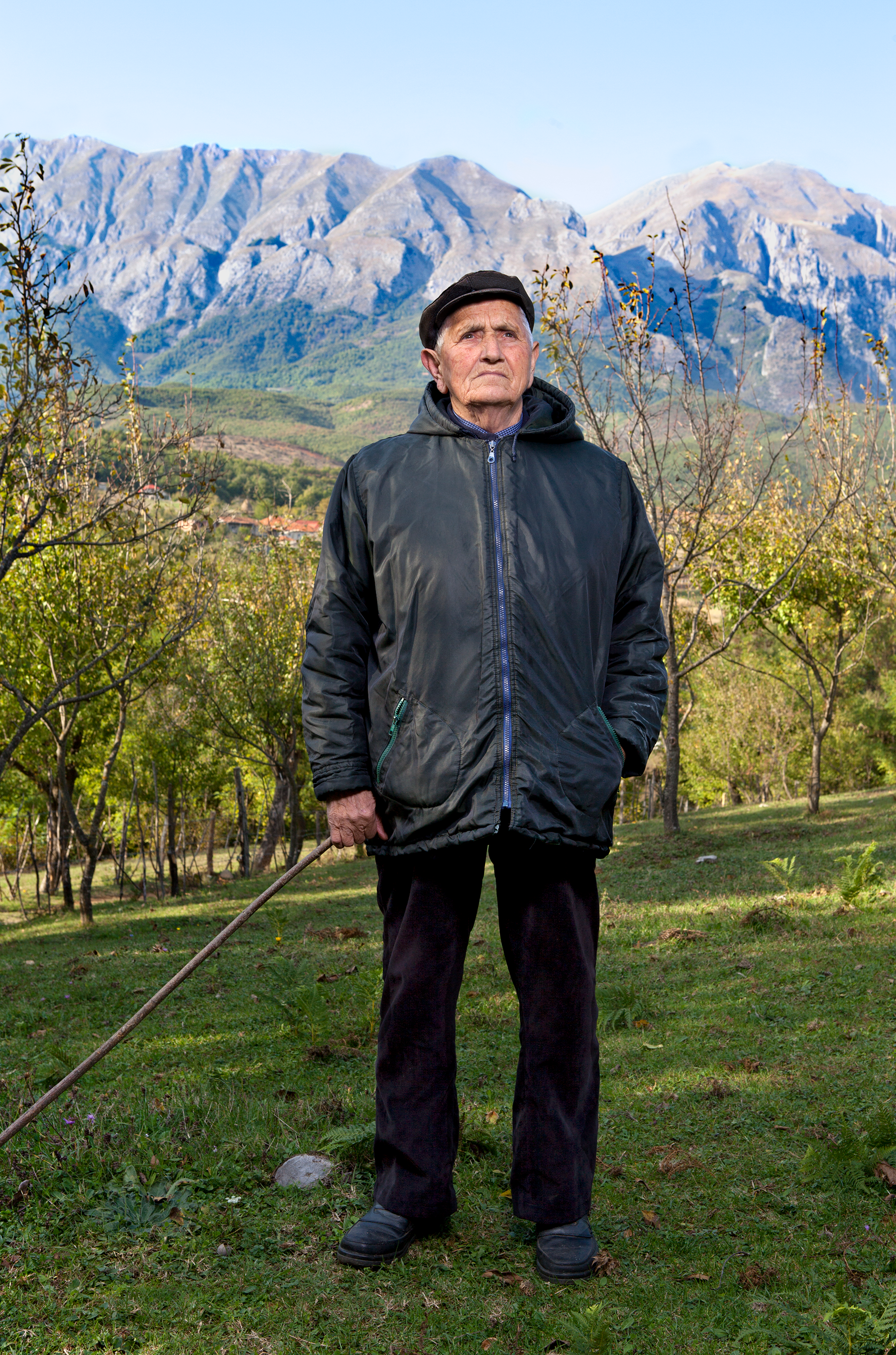 A person in a large black raincoat and black slacks looks up in front of an orchard.