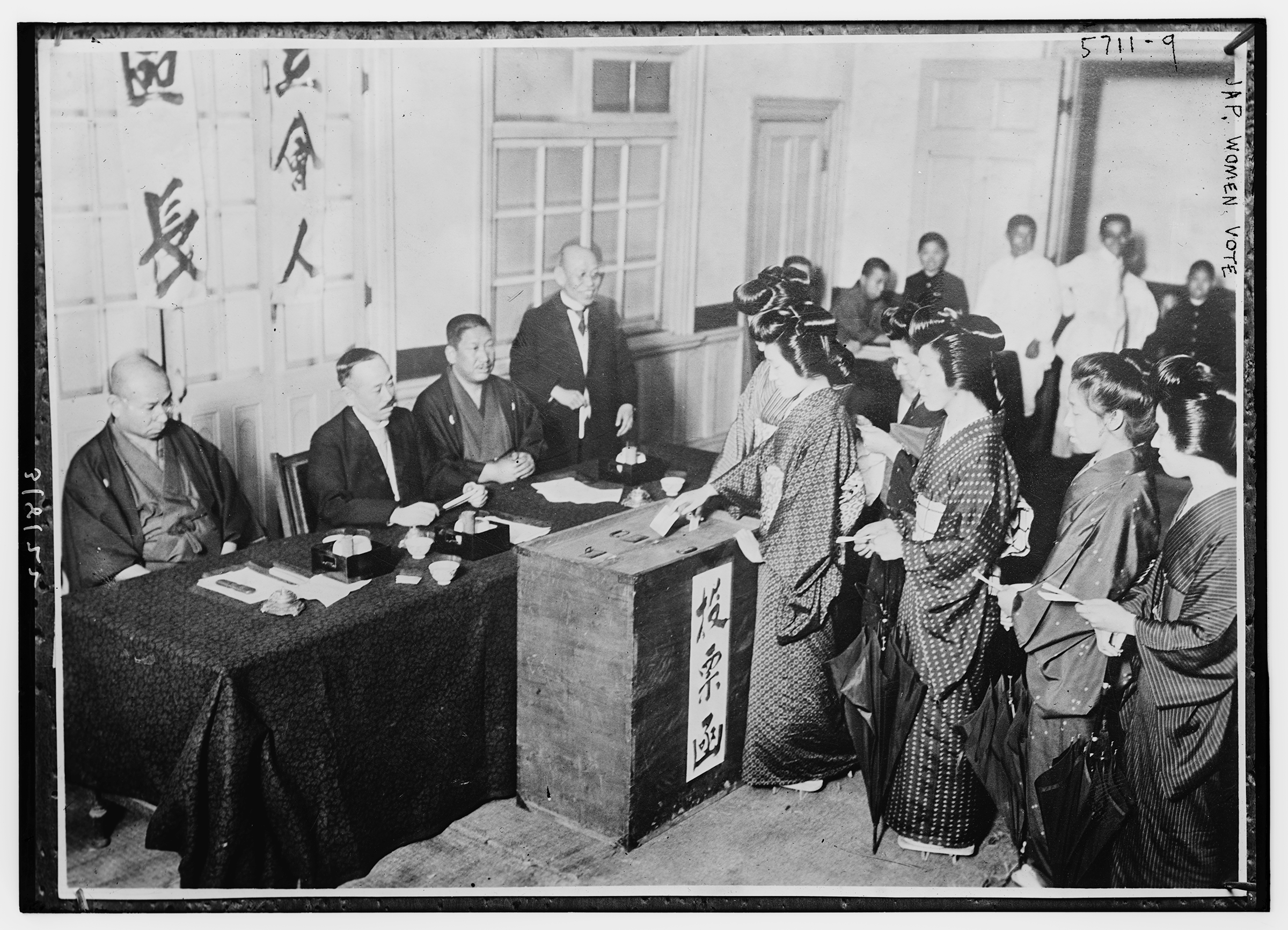 A black-and-white photo of a line of Japanese woman putting slips into a large box in front of a table of Japanese men.