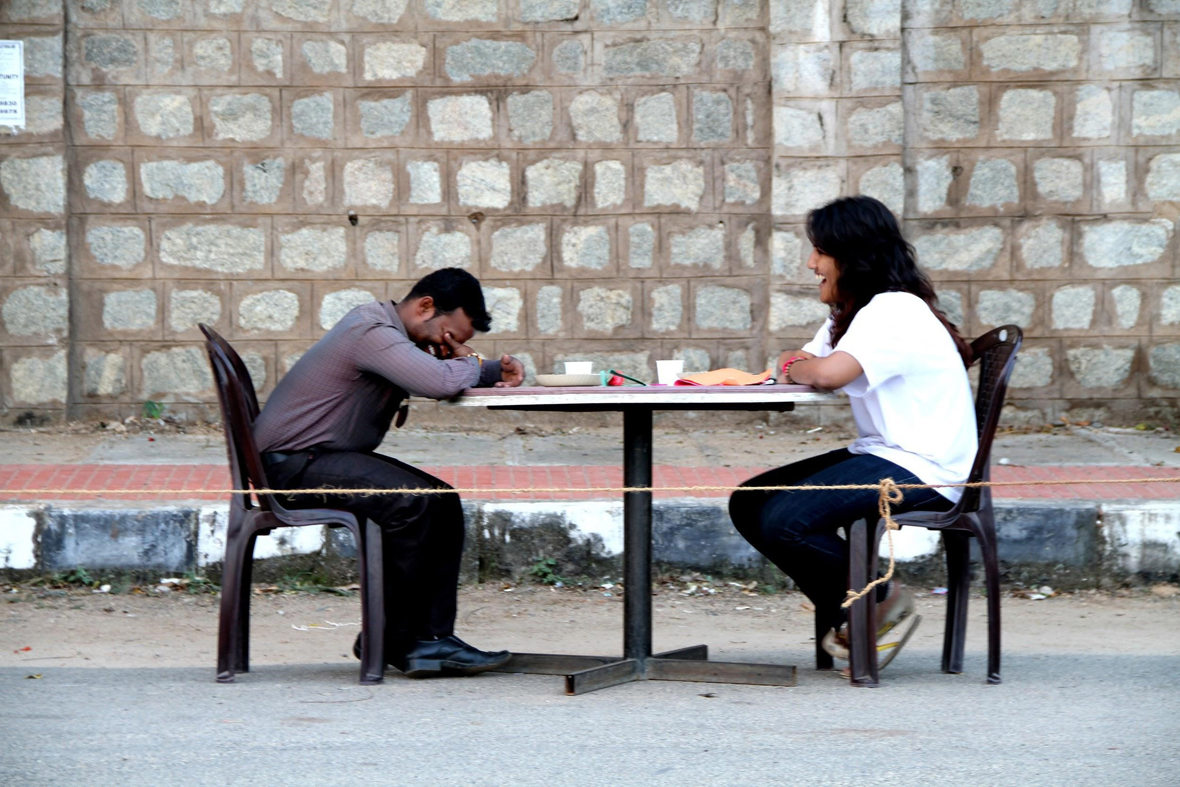A man and a woman sit across from each other at a table outdoors, smiling.