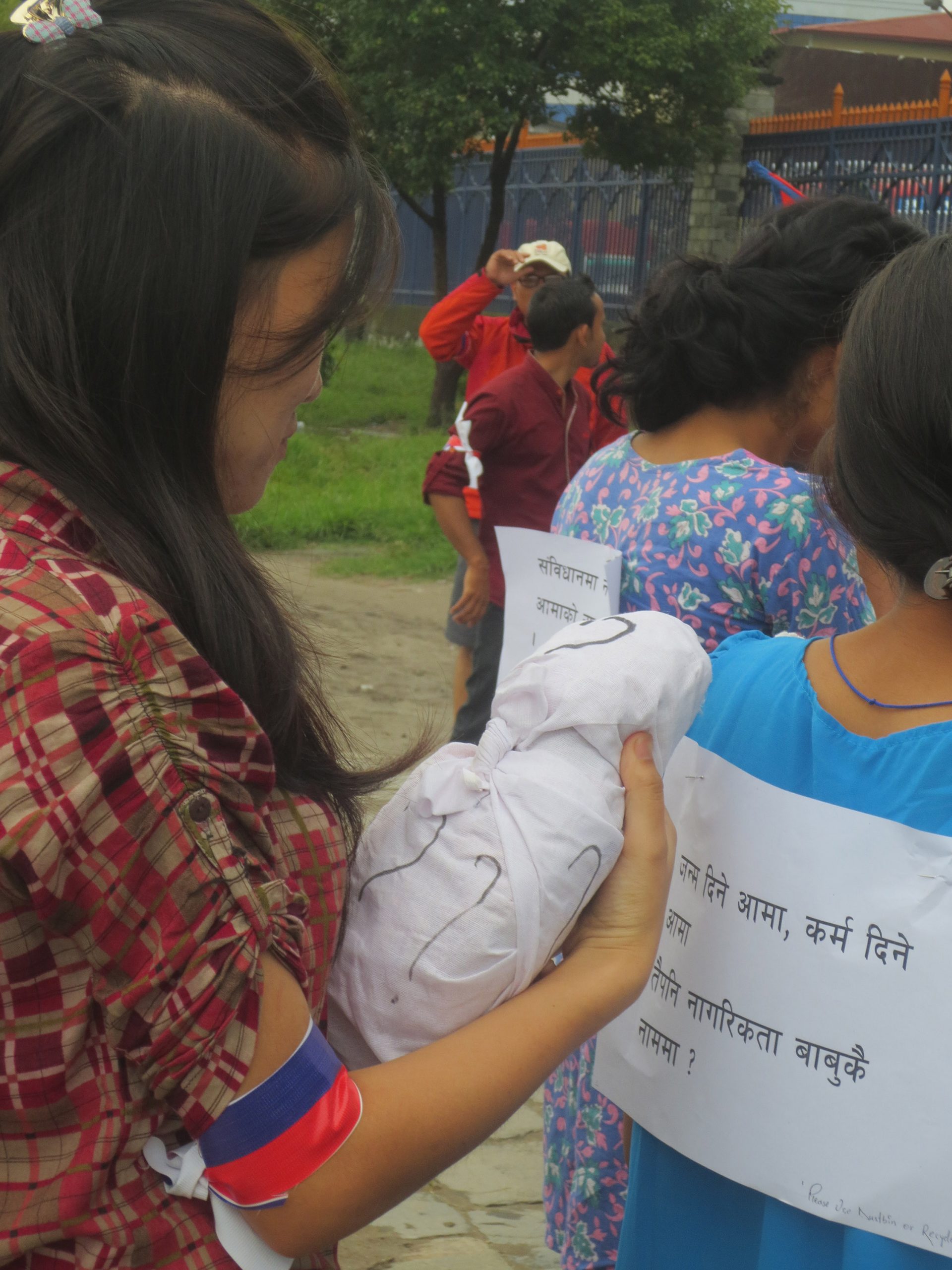 A young woman holds the white-wrapped effigy of a baby covered in question marks, with protestors in front of her with signs pinned to their backs.