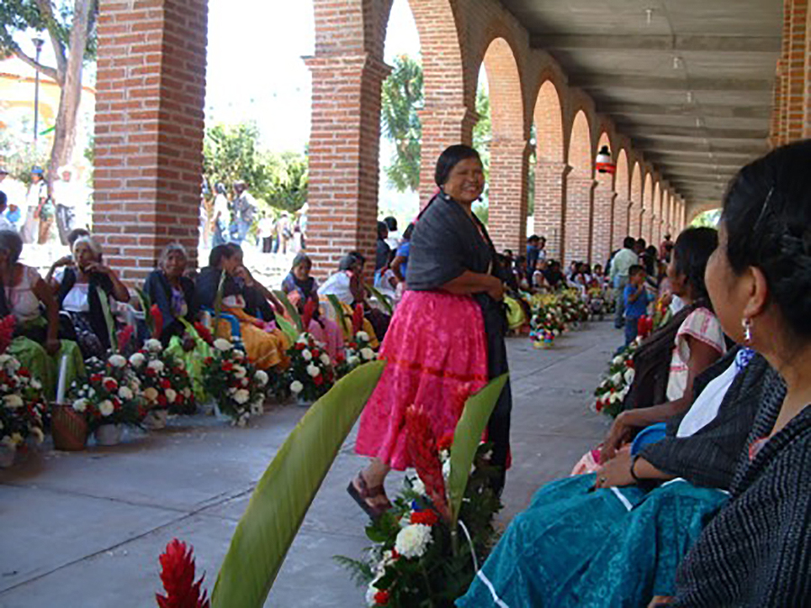 A woman is standing and smiling in an open corridor, women sitting along the sides behind elaborate flower bouquets.