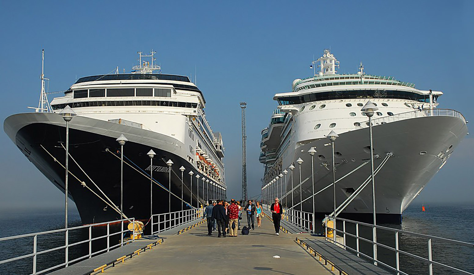Two large cruise ships docked side-by-side with a group of white tourists between them.