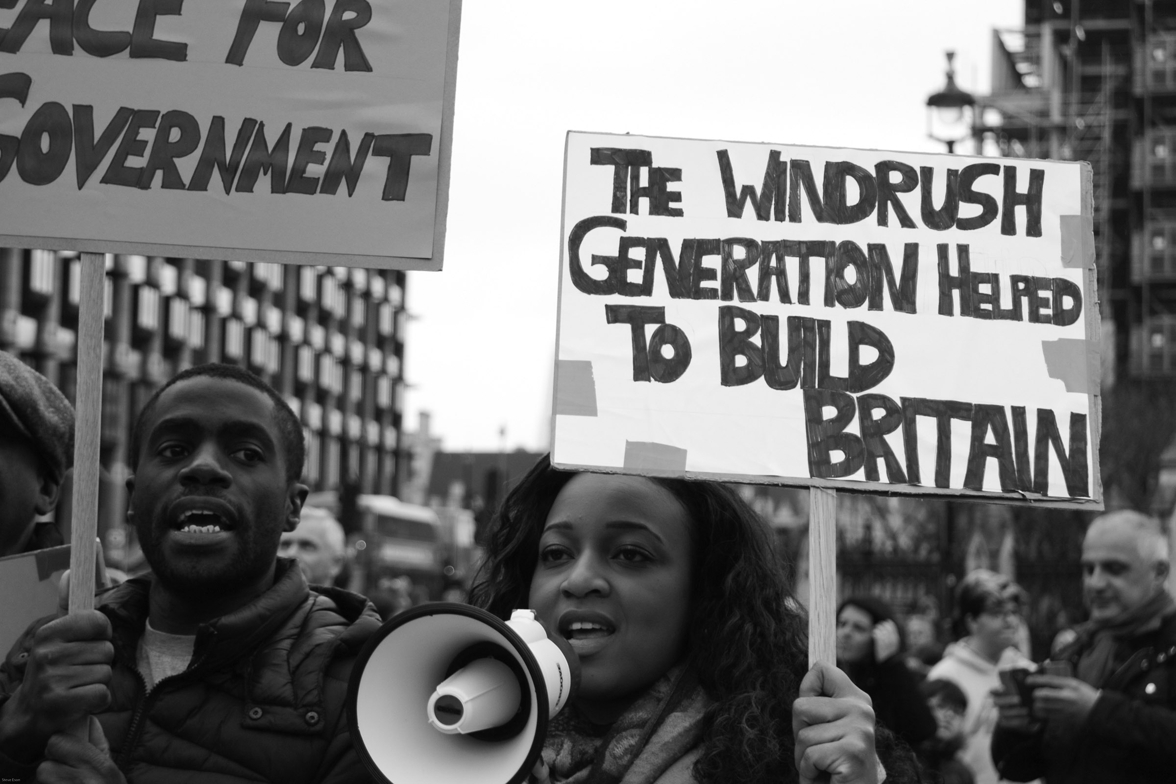 Two people, one with a megaphone, hold signs on a street; one sign says "The Windbrush Generation helped to build Britain."