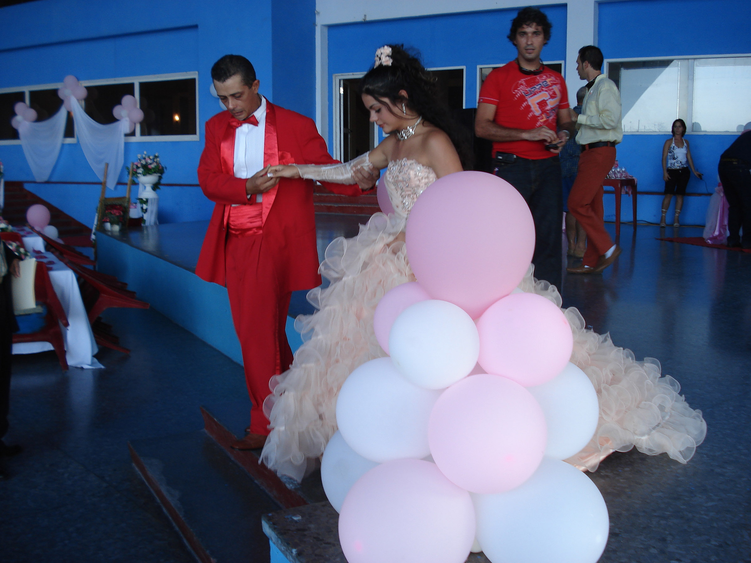 A young girl in a lavish light pink dress is led to the dance floor by a man in a red suit.