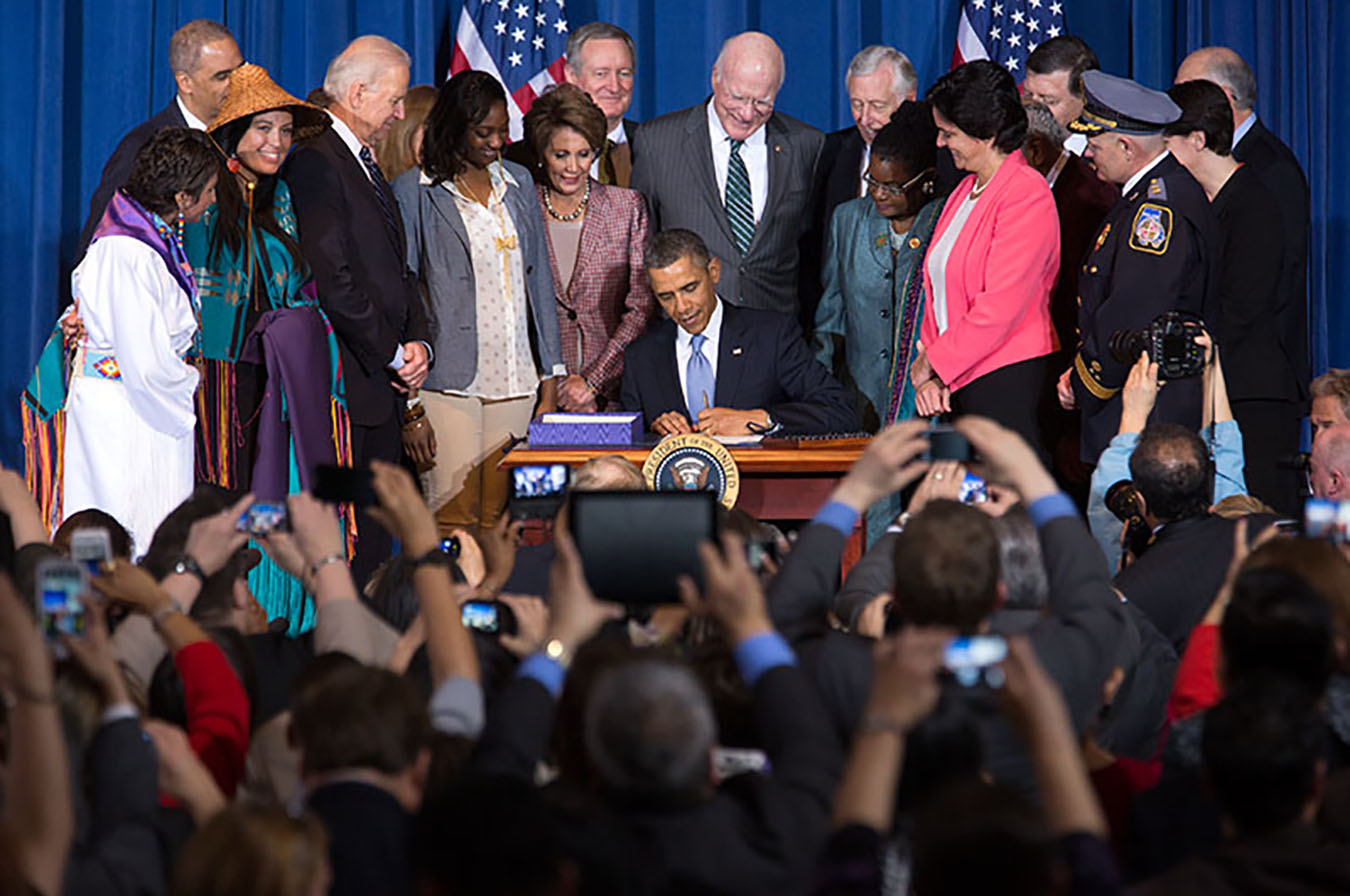 U.S. President Barack Obama signs a document in front a group of photographers and surrounded by a group of officials and activists.