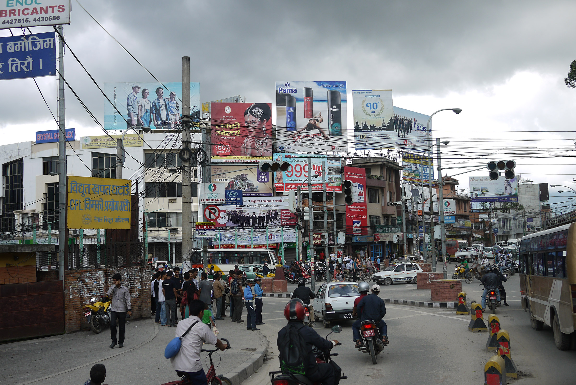 A very busy street in Kathmandu; there are people on the sidewalk and driving cars and bikes, billboards in the distance.
