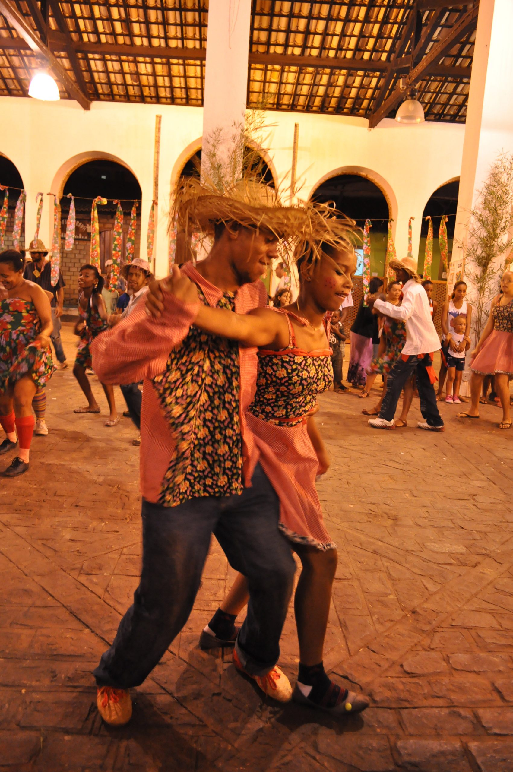 A young man and woman dance, the man wearing a straw hat, at night under bright lights.
