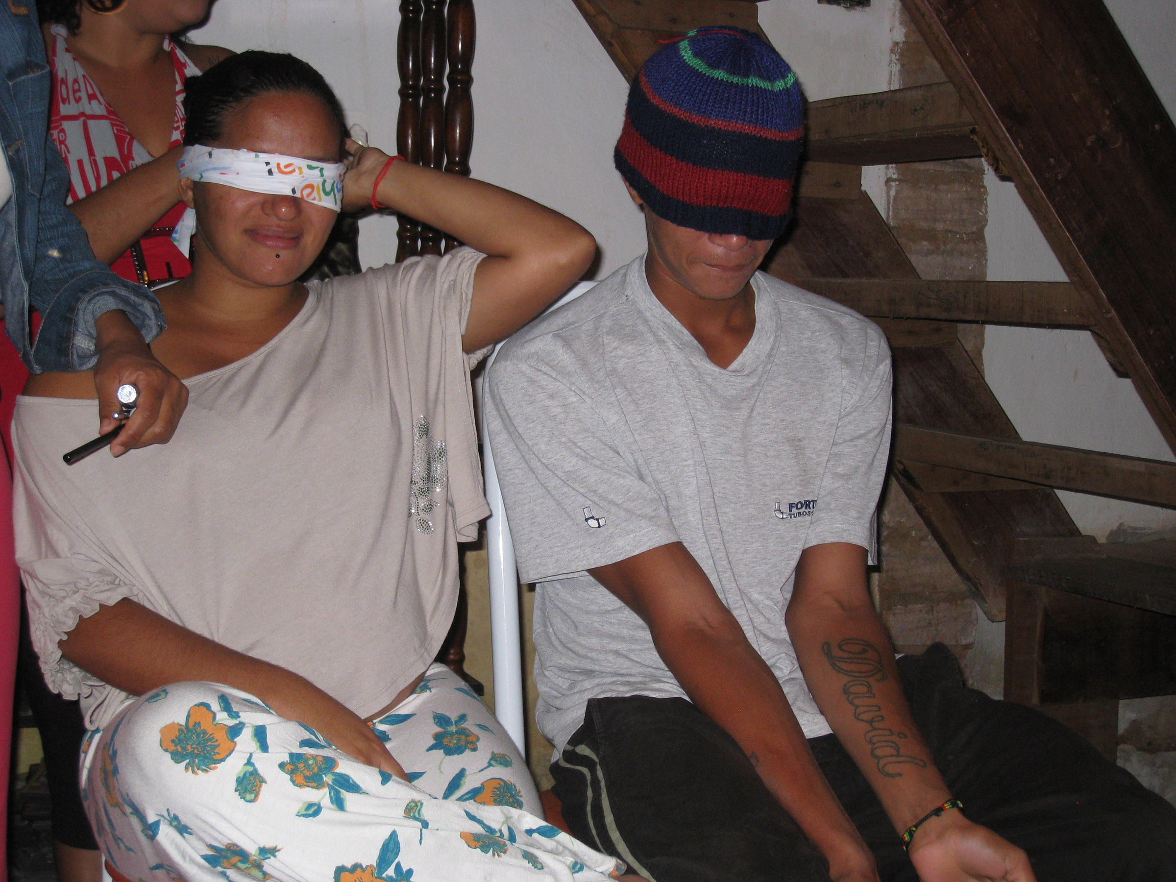 A young man and woman sit next to each other, the man with a hat over his eyes and the woman a blindfold.