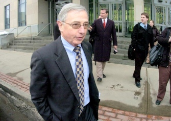 A man in a suit and tie leaves a courthouse, press following him.