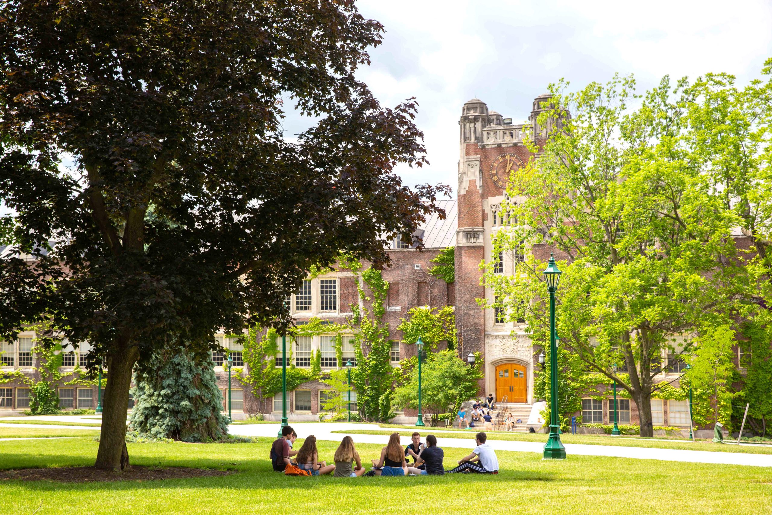 College students sit in a circle on the ground in the shade of a tree. Behind them is a brick building with a clock tower and a lamp post.