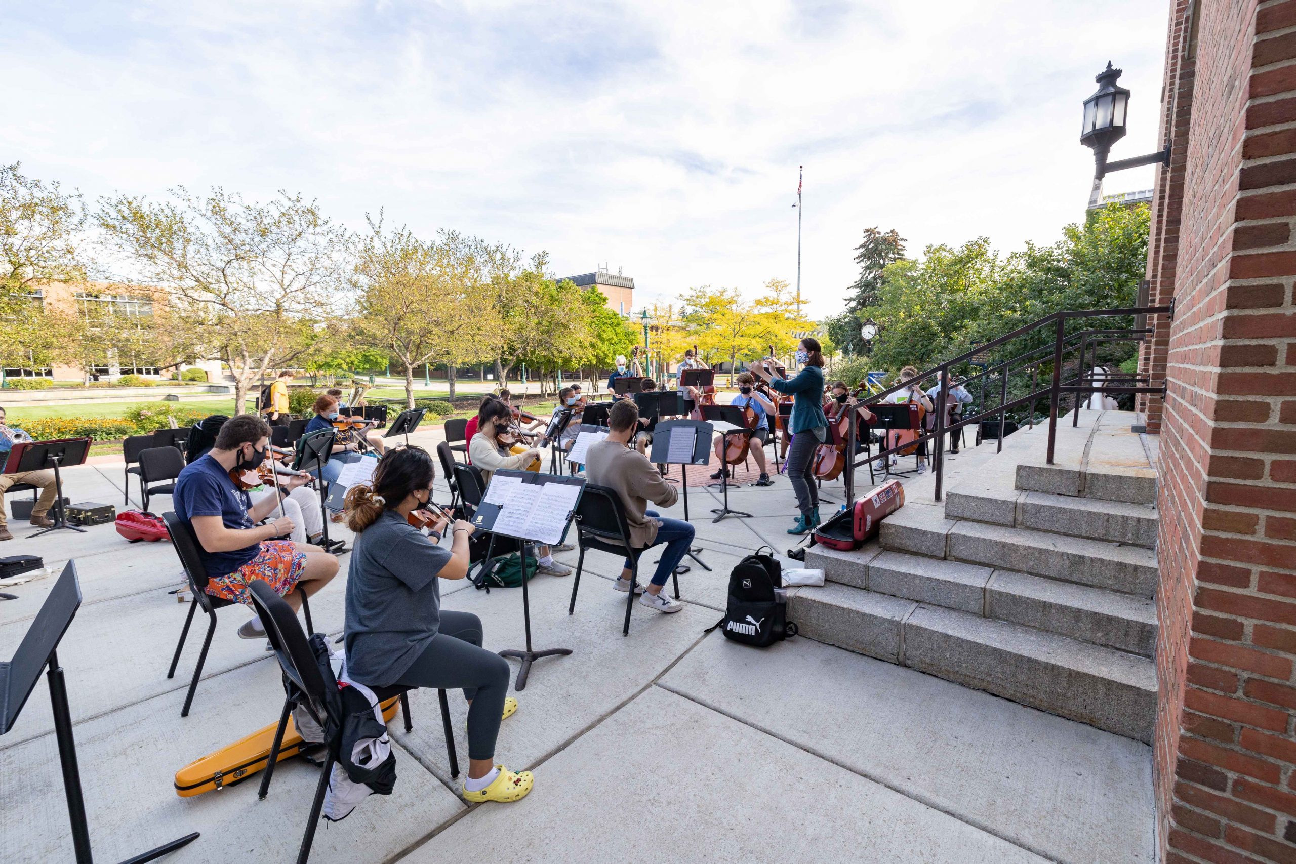 A string orchestra practices outside on a sidewalk near a campus quad.