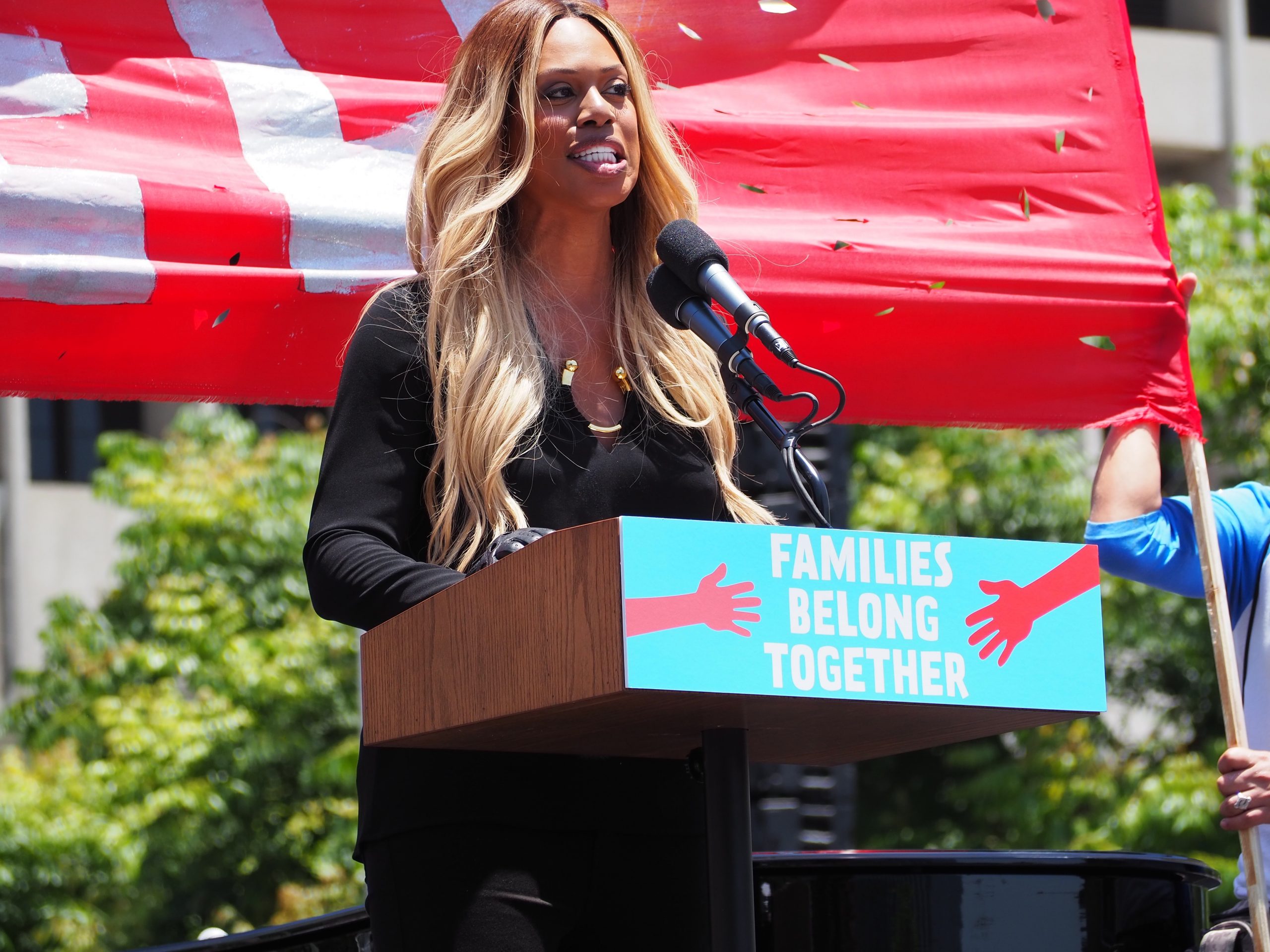 A woman speaks from a microphone in front of the sign "FAMILIES BELONG TOGETHER."