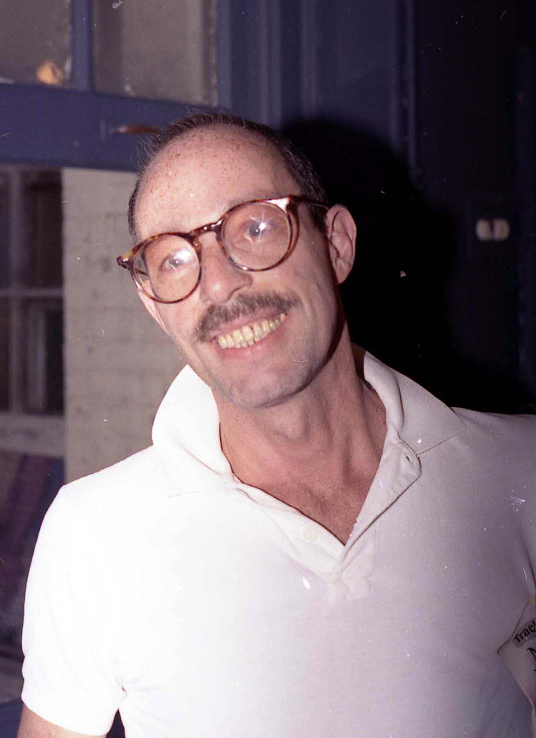 A man in a white shirt and large glasses smiles at the camera.