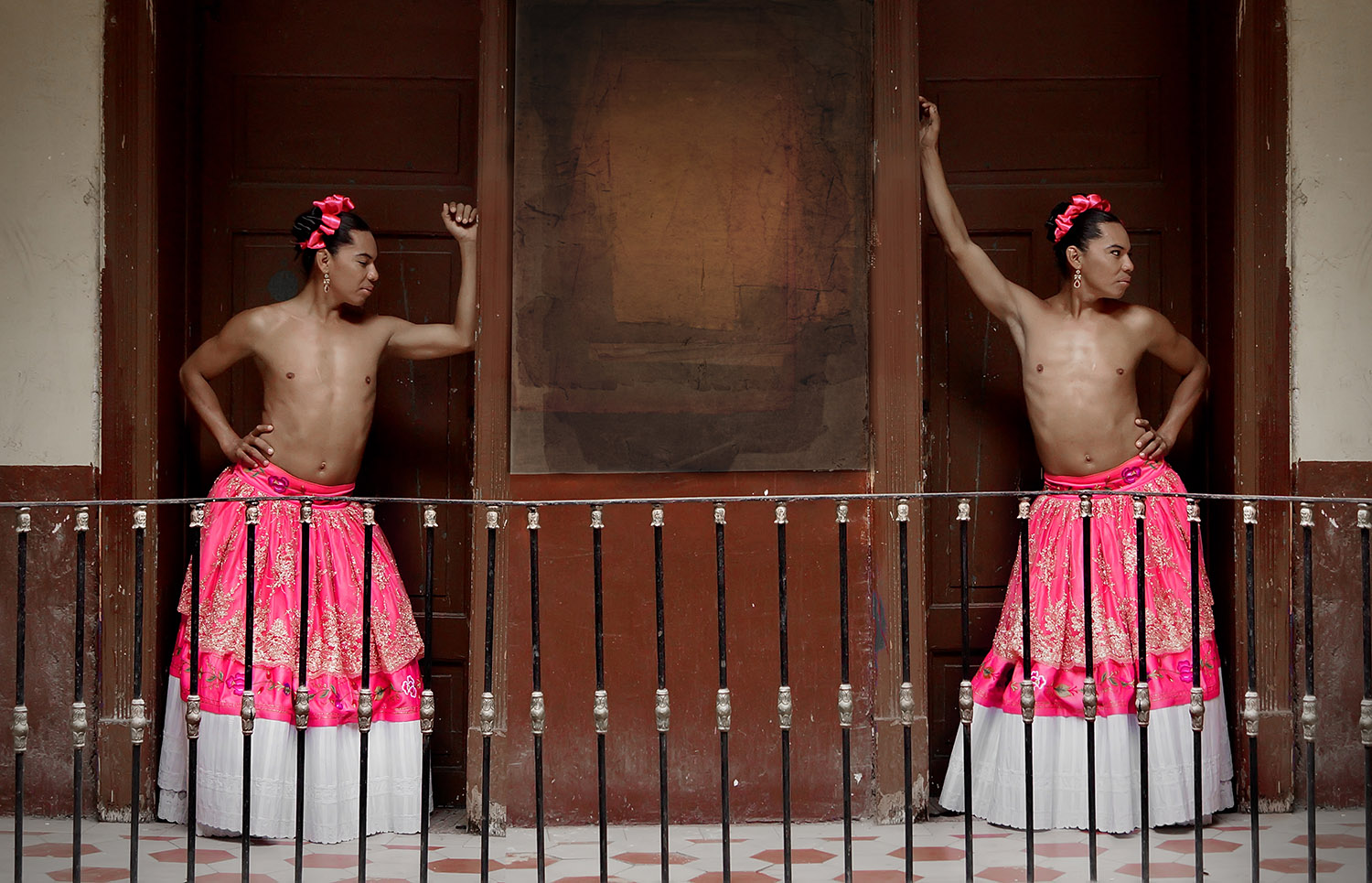 Two identical people lean against a doorframe, shirtless wearing a pink skirt and pink bow.