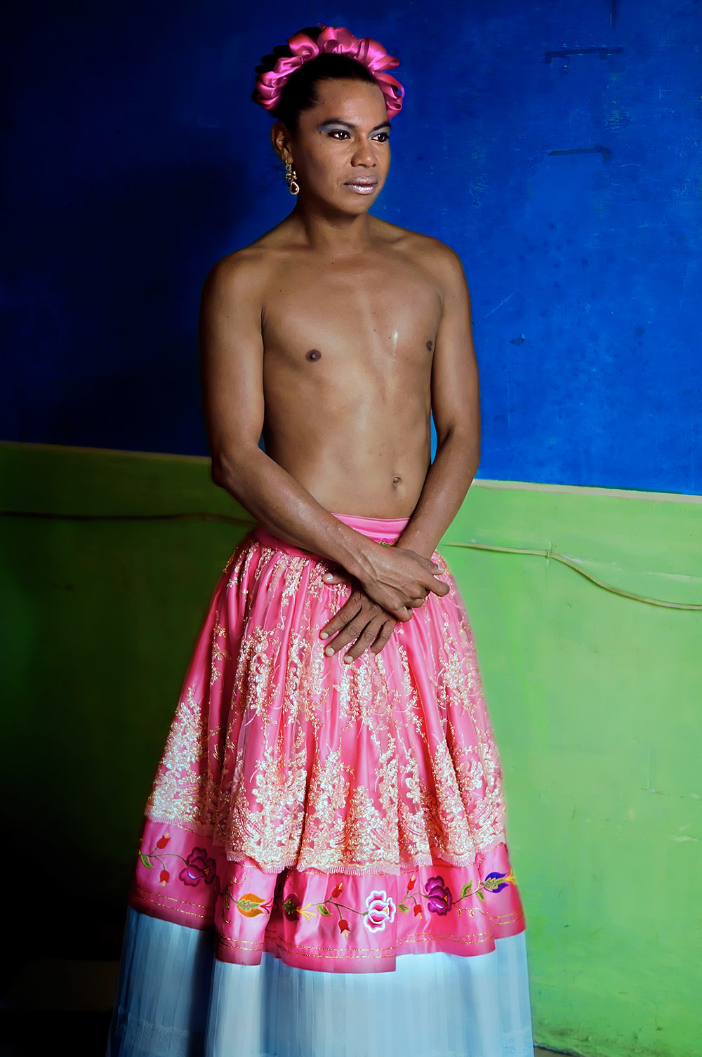 A shirtless muxe wearing a pink skirt and make-up looks off-camera.