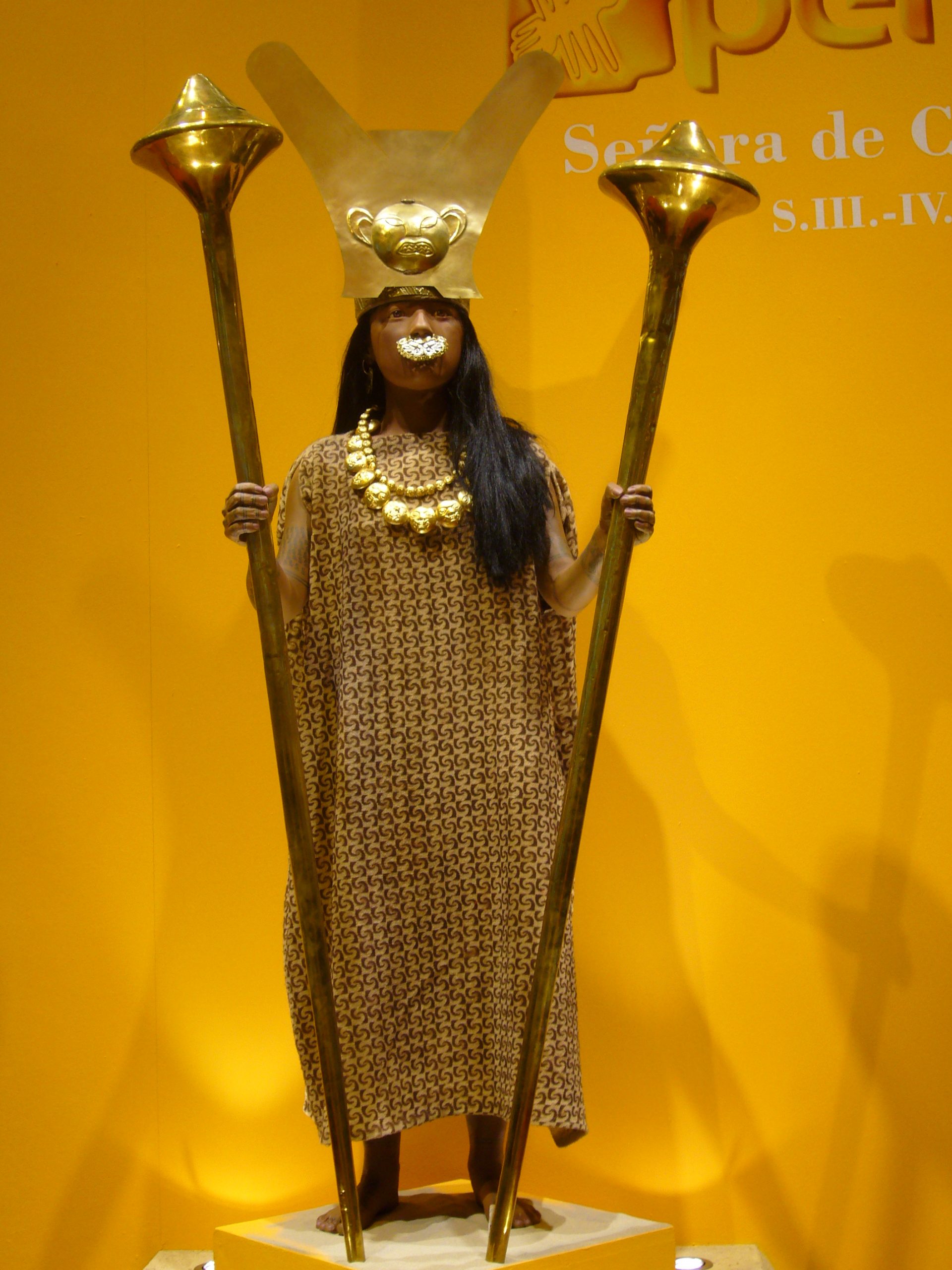 Museum display of a woman with elaborate gold jewelry and large gold staves.