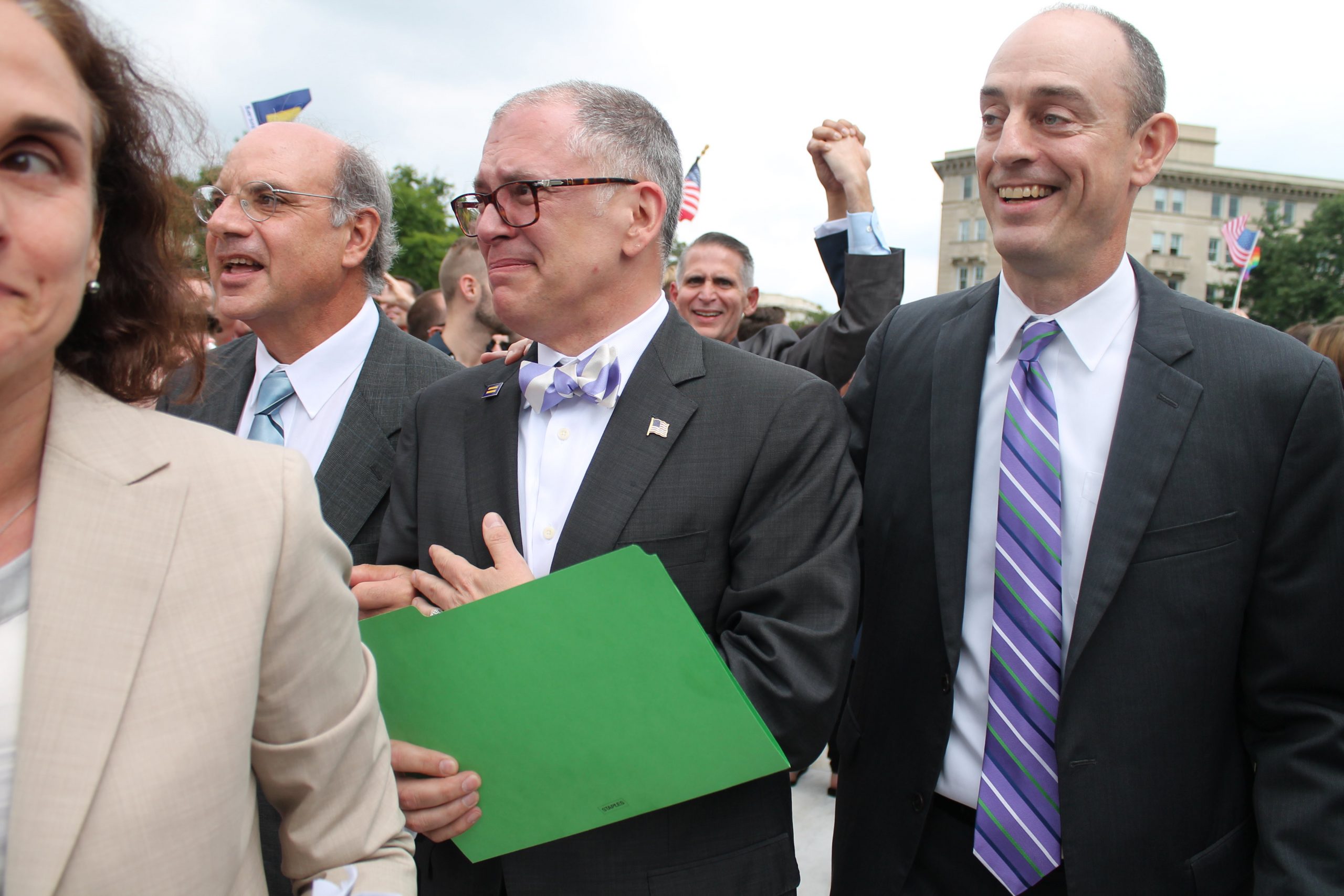 A man is emotional holding a green folder, two others smile around him.