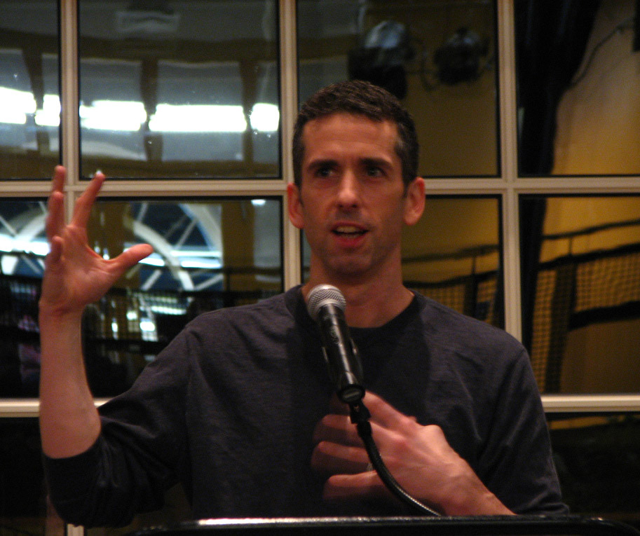 A man speaks in front of a microphone, one hand raised.