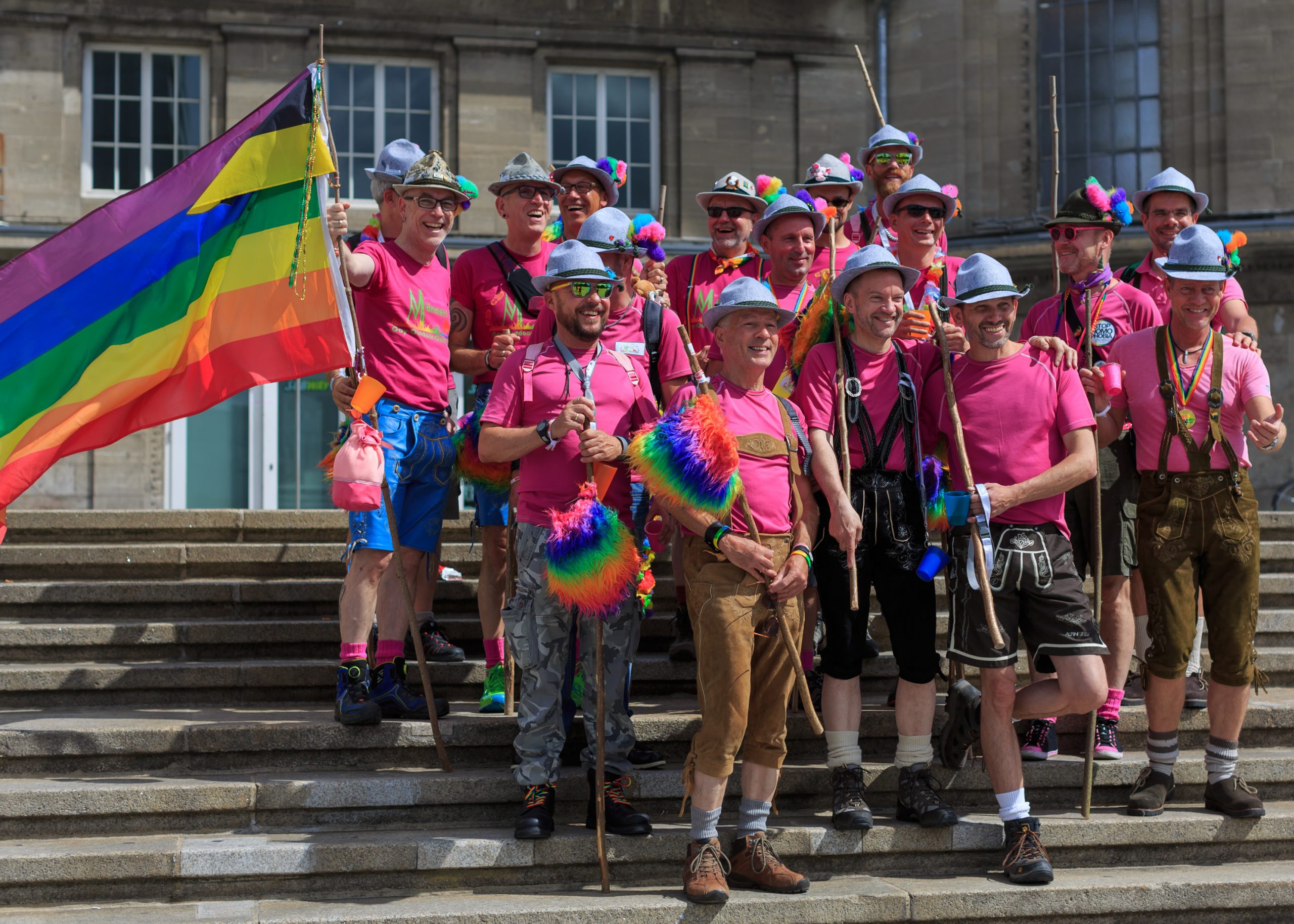 A group of men in pink shirts hold rainbow flags with walking sticks.