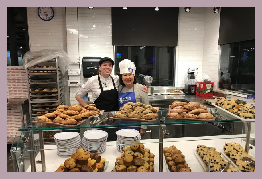 Two people smiling proudly in front of multiple platters of cookies, muffins, and scones.