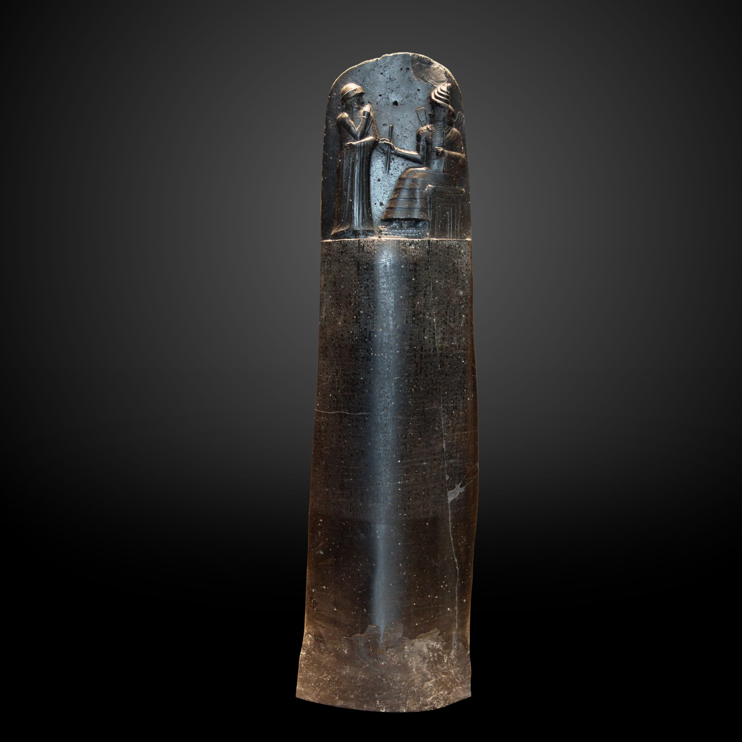 Tall, black stone stele with a carved top section showing two figures, one seated. It is positioned on a display in a museum setting.