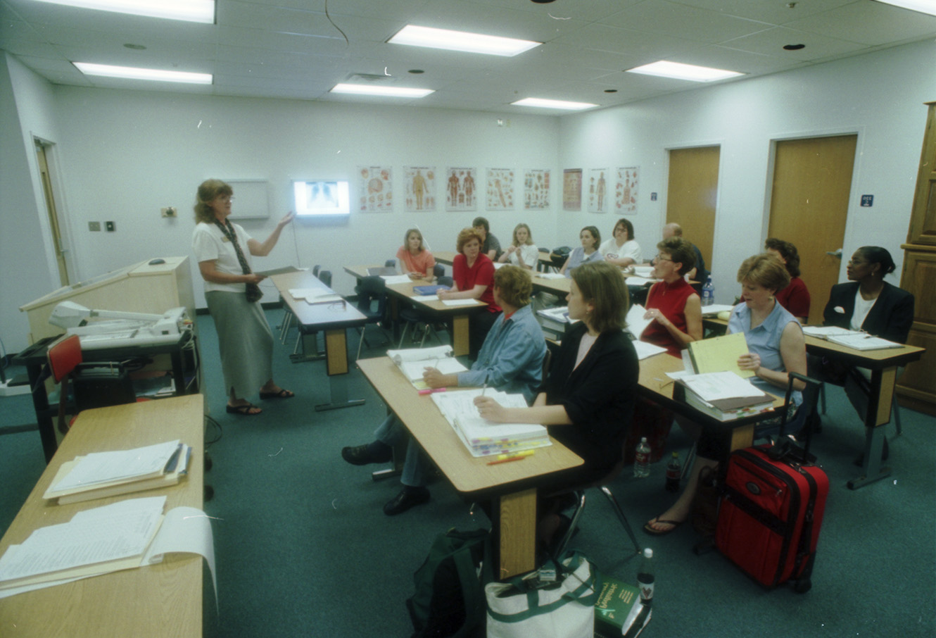 College of Nursing classroom scene—female teacher addressing adult students, who are seated at desks with papers. Educational posters line the walls.