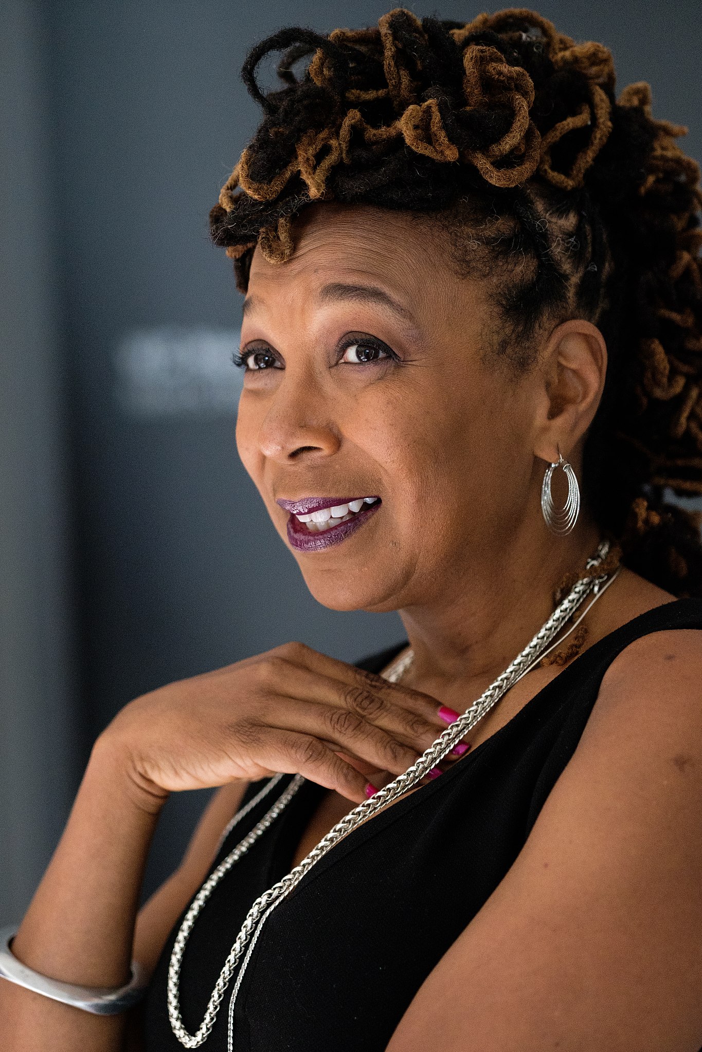 A woman with styled locs and silver hoop earrings smiles, wearing a sleeveless black top and silver necklace.