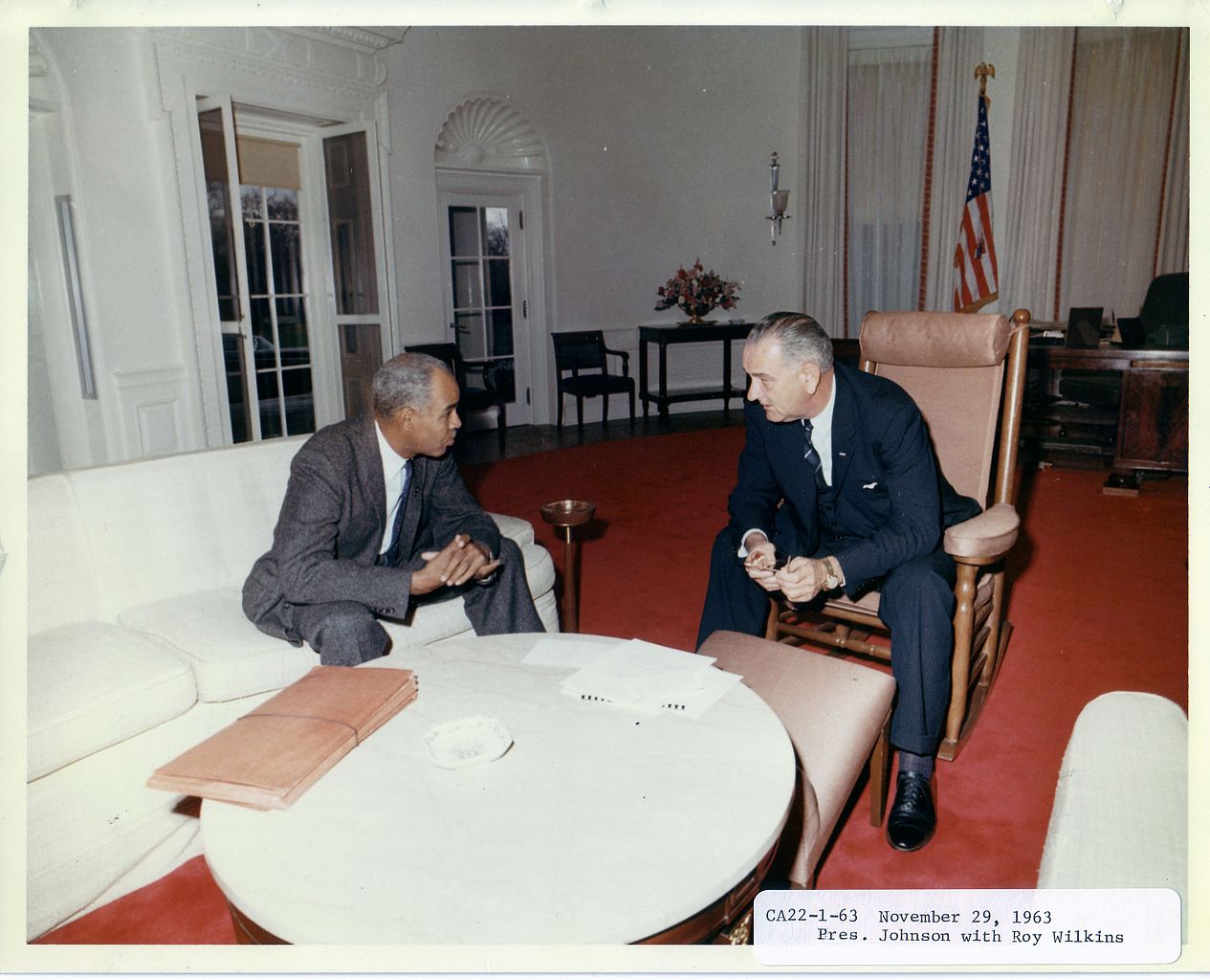 Two men sit conversing in a formal office with a red carpet and U.S. flag.