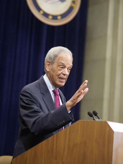 A man in a suit and tie gestures while speaking at a podium in a formal setting. A seal hangs on a dark curtain behind him.