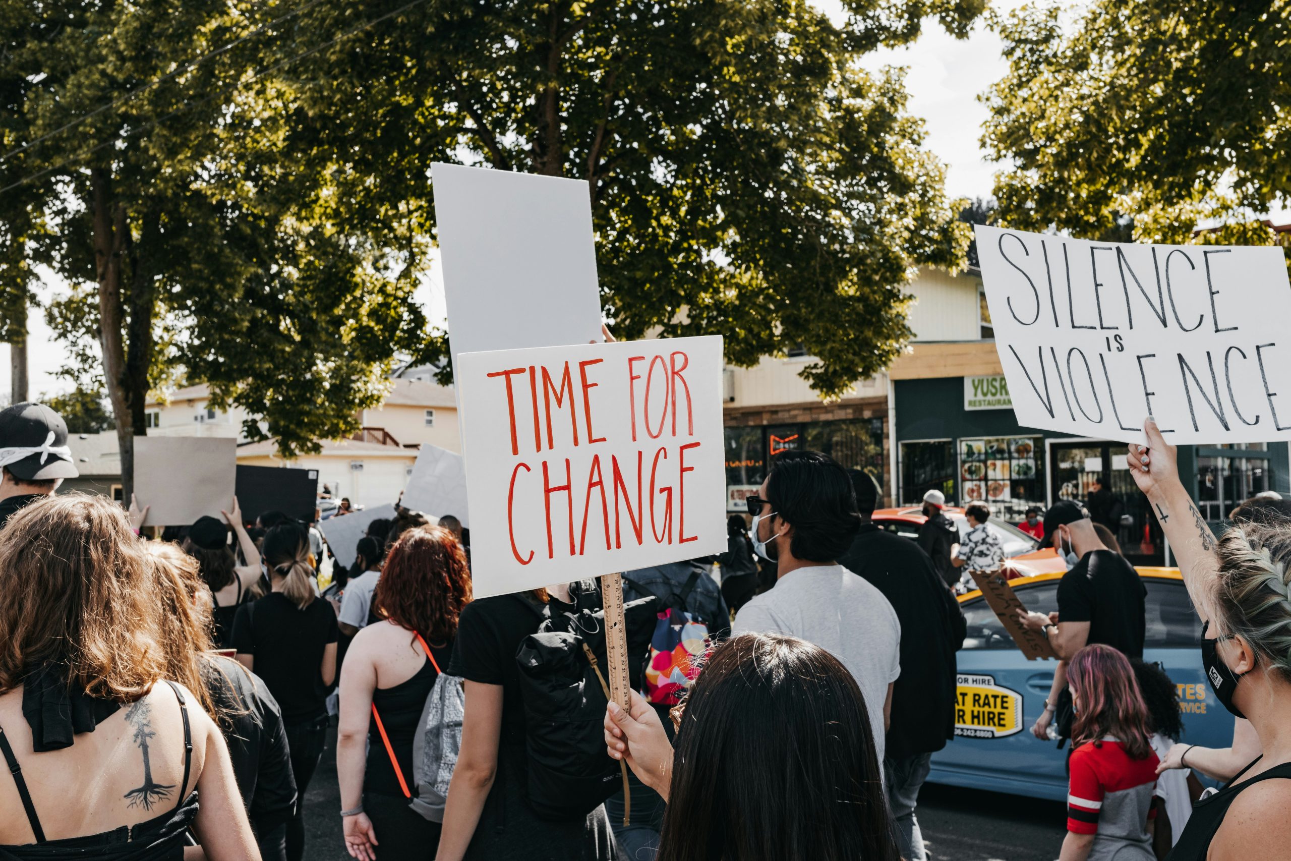 A group of diverse protesters march under trees, holding signs reading "TIME FOR CHANGE" and "SILENCE IS VIOLENCE,"