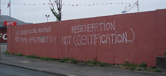A red wall with white graffiti reads, "We need social housing, not yuppy apartments... Regeneration not (gentrification...)."