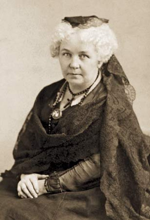 Sepia-toned portrait of Elizabeth Stanton. She has curly, white hair, is wearing a dark lace shawl and ornate necklace. She has a serious, thoughtful expression.
