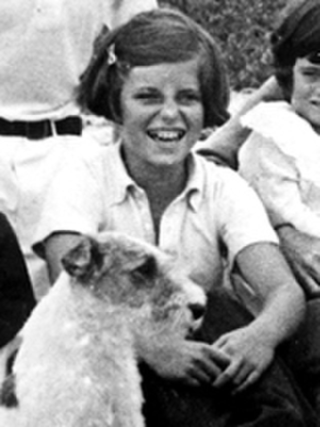 Cropped black and white photo of a young girl with a short bob hairstyle and a clip, smiling joyfully. She is wearing a collared shirt and is sitting next to a fluffy dog.