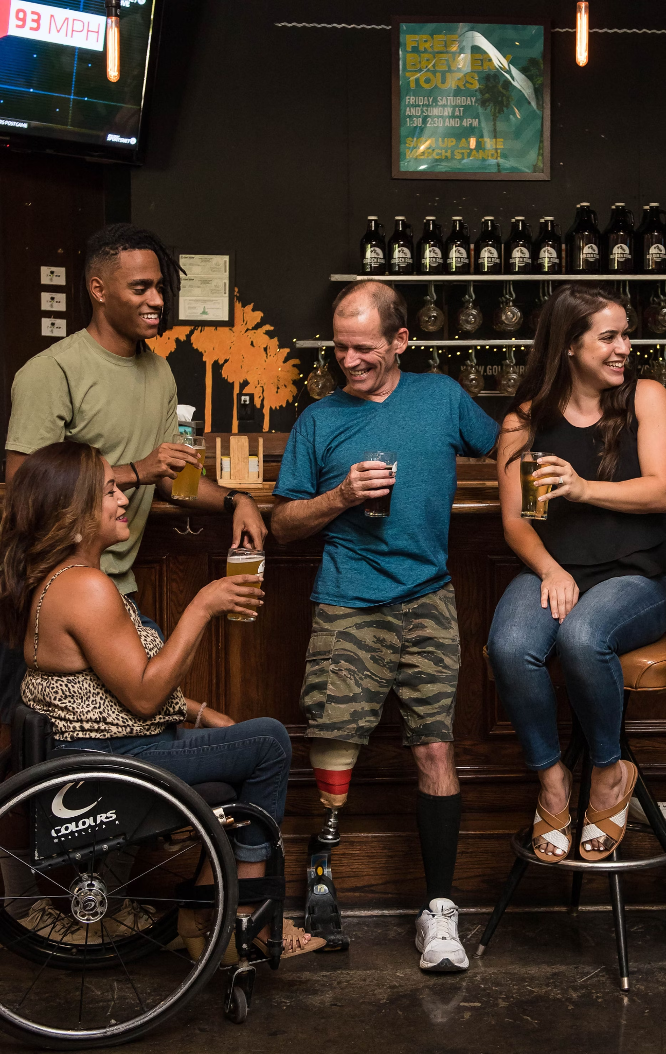 A diverse group of four friends enjoy drinks at a bar. They are smiling and chatting, creating a warm, inclusive atmosphere. One person is in a wheelchair, and another has a prosthetic leg, highlighting diversity and accessibility.