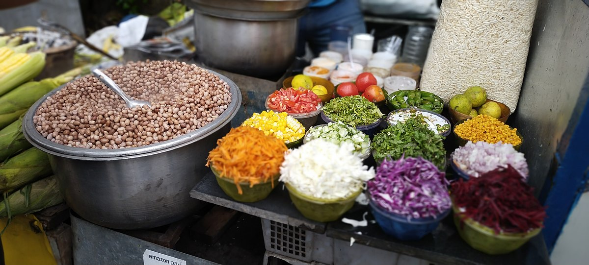Variety of fresh vegetables and grains in bowls on a street food stall, including peanuts, lettuce, cabbage, and spices, conveying a vibrant, colorful scene.