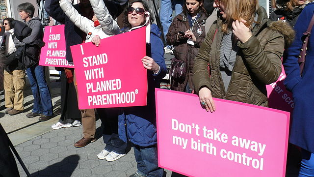 Protesters hold bright pink signs reading "I Stand with Planned Parenthood" and "Don't take away my birth control."