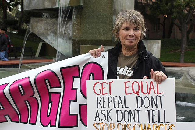 A person stands holding a protest sign advocating to repeal "Don't Ask, Don't Tell" in front of a fountain.