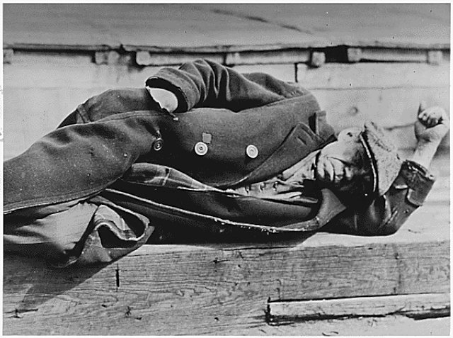 An old black and white photograph of an older man in a worn heavy coat and flat cap sleeping on a wooden dock. His expression is wearied, conveying exhaustion.