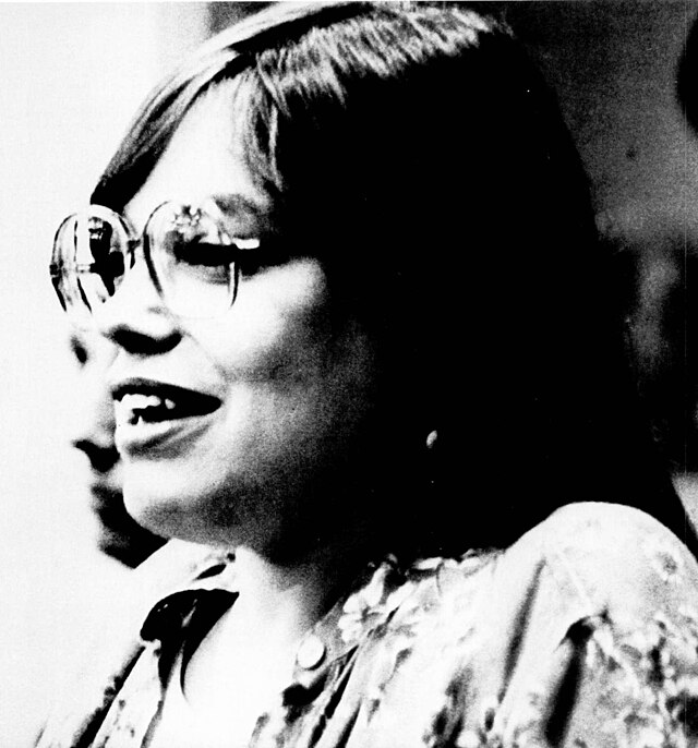 A portrait of a woman with glasses shown from the side. She smiles softly, her gaze slightly upward. She's wearing a patterned blouse, and the image has a grainy black and white effect.