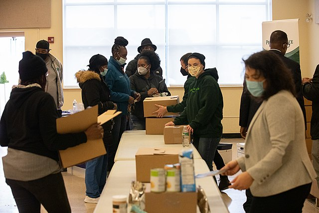 A diverse group of people, wearing masks, are distributing and receiving food boxes in a community center.