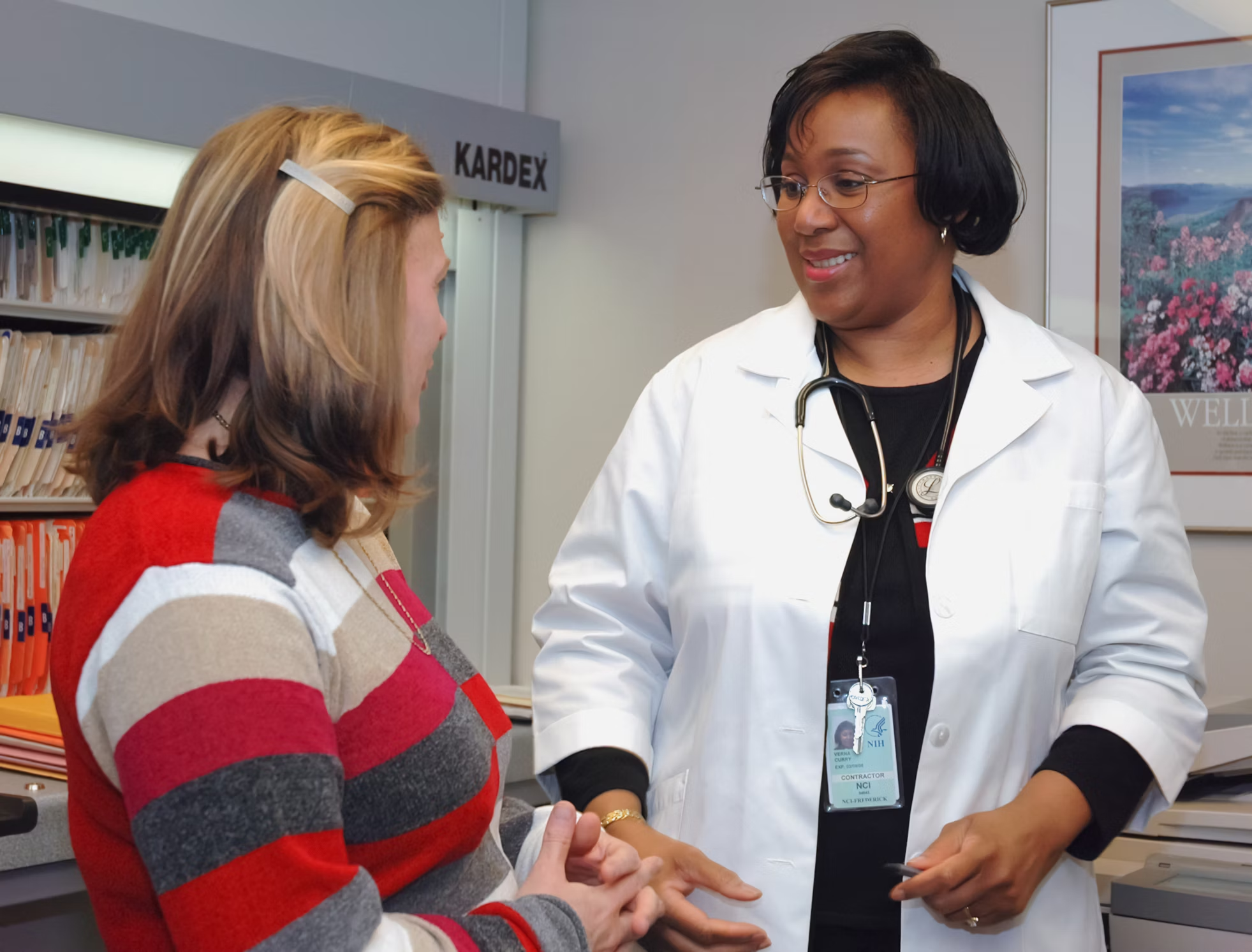A doctor in a white coat with a stethoscope smiles while conversing with a woman in a striped sweater. They stand in an office with medical files.
