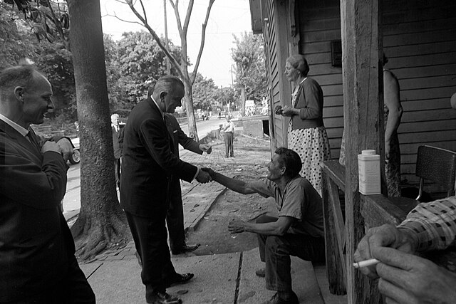 An old black and white photo. The president shakes hands with a seated man on a porch, surrounded by others. Trees and cars line the road.