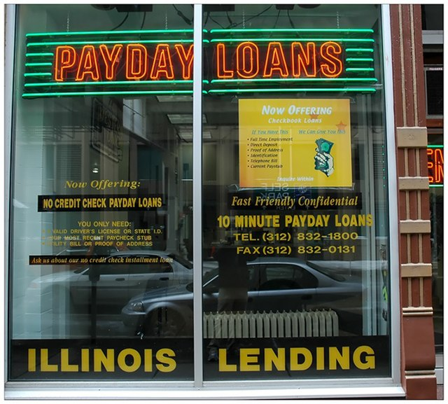 Storefront window advertising payday loans in bright neon signs with yellow and red text. Promises fast, no credit check loans. Urban street reflection.