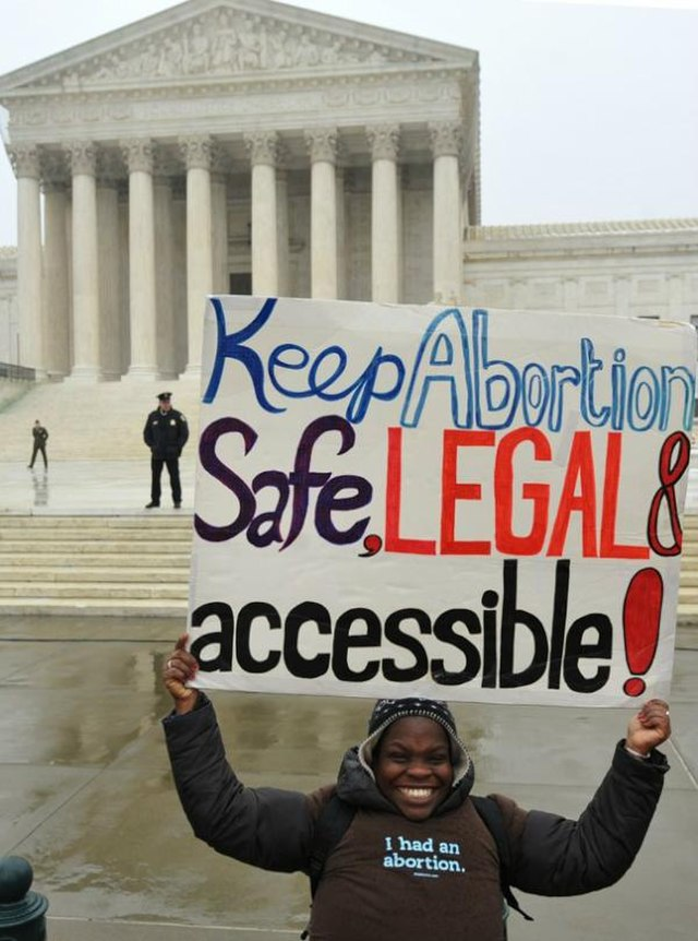 A person smiles while holding a sign reading "Keep Abortion Safe, Legal & Accessible!" in front of the U.S. Supreme Court.