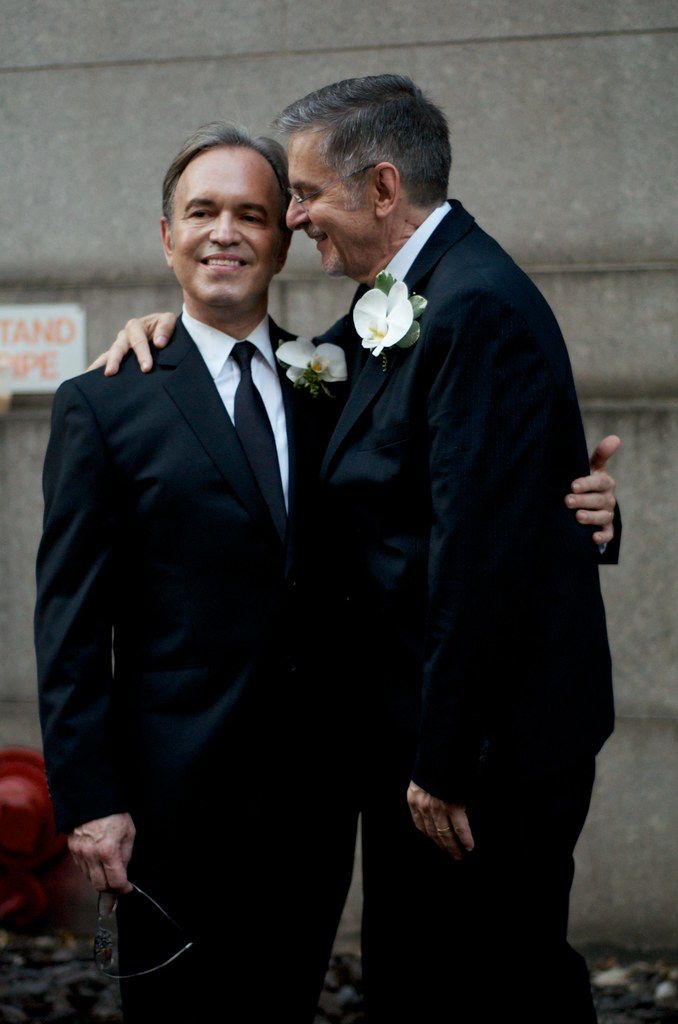Two grooms in black suits embrace affectionately, each wearing a white flower on their lapel, conveying warmth and joy against a stone wall backdrop.