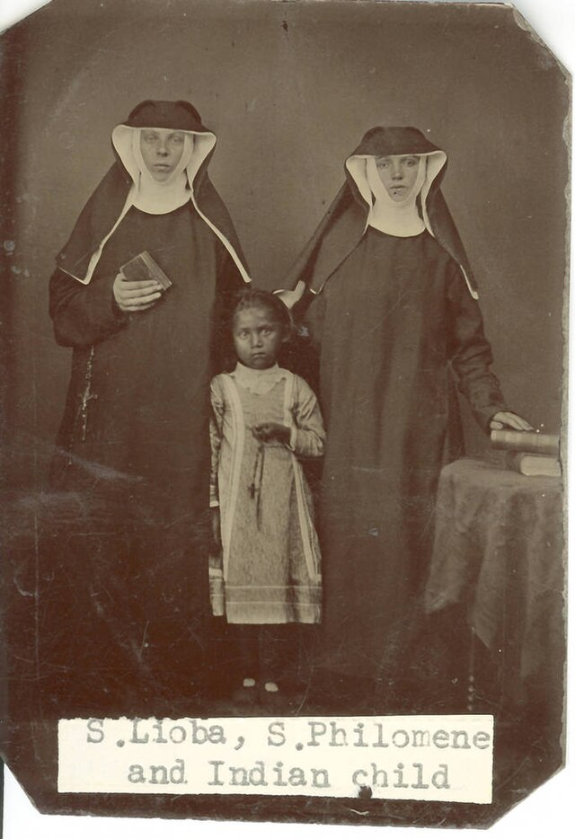 Two nuns in traditional habits stand beside a young girl. The nun on the left holds a book, while the other rests a hand on a table. The girl looks forward solemnly. A typed label reads, “S. Lioba, S. Philomene and Indian child.”