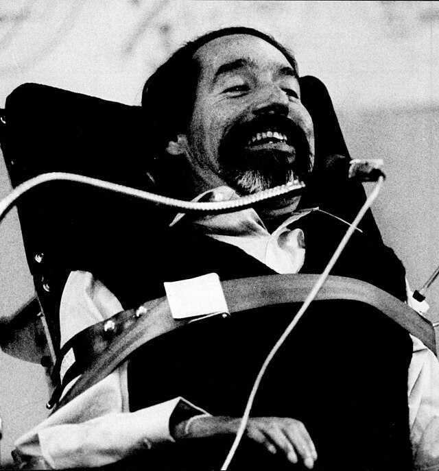 Smiling man in a wheelchair using a mouth-controlled device. He wears a dark vest and white shirt. Black and white photo.