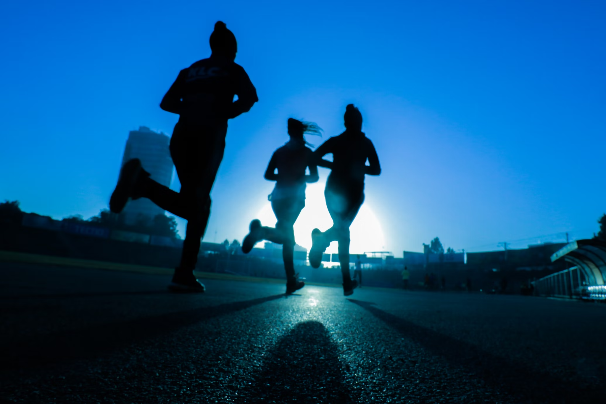 Silhouettes of three people running on a track with a bright sun setting behind them, casting long shadows. The scene conveys energy and motion.